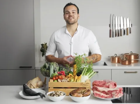 Jules Cooking in his kitchen behind a crate of vegetables, a whole fish, shellfish and beef